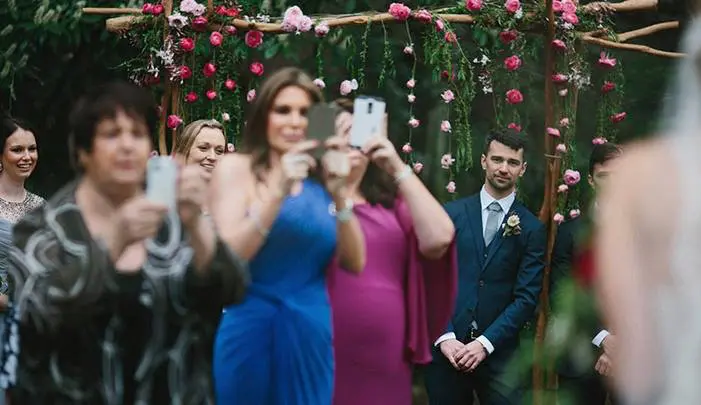 Guests filming the ceremony on their smartphones in front of the flower arch where the groom is waiting