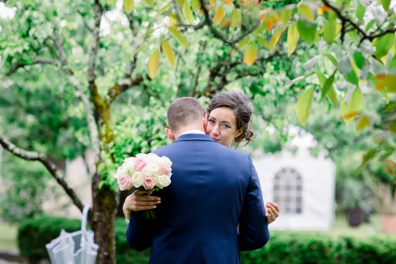 Groom hugging the bride beneath a garden tree, rose bouquet in hand