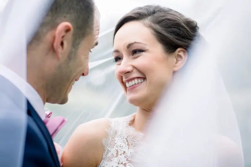 Bride laughing under her veil facing the groom in a blue suit