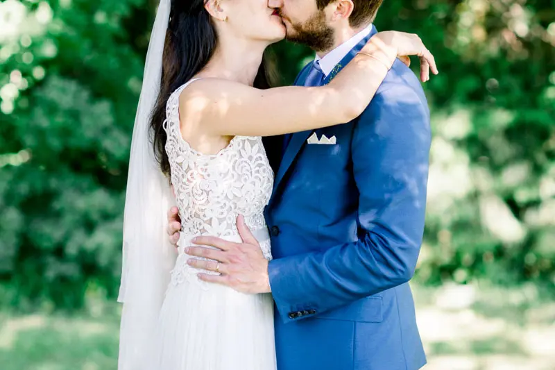 Bride and groom kissing in the greenery, lace gown and blue suit