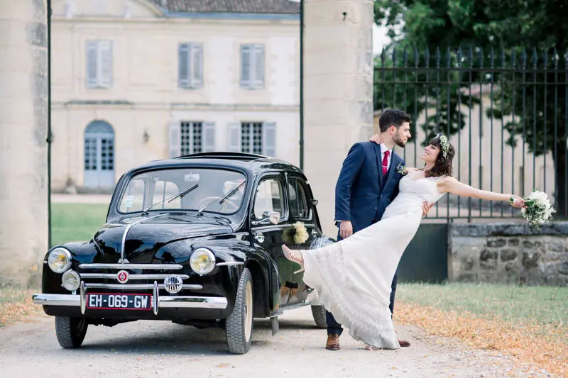 Bride posing playfully by the vintage car in front of Château Goudichaud