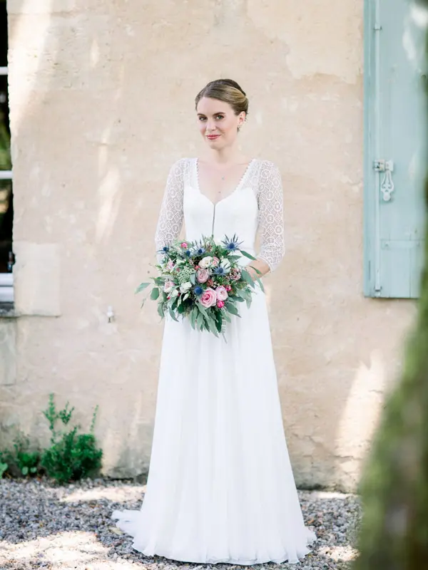 Bride in a lace gown holding her colourful bouquet in front of a pale wall