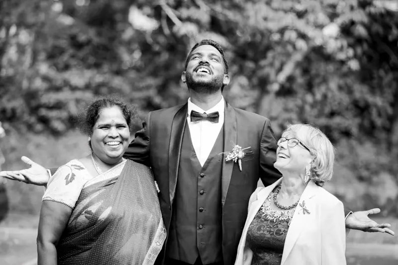 Groom flanked by two joyful women with open arms, black and white