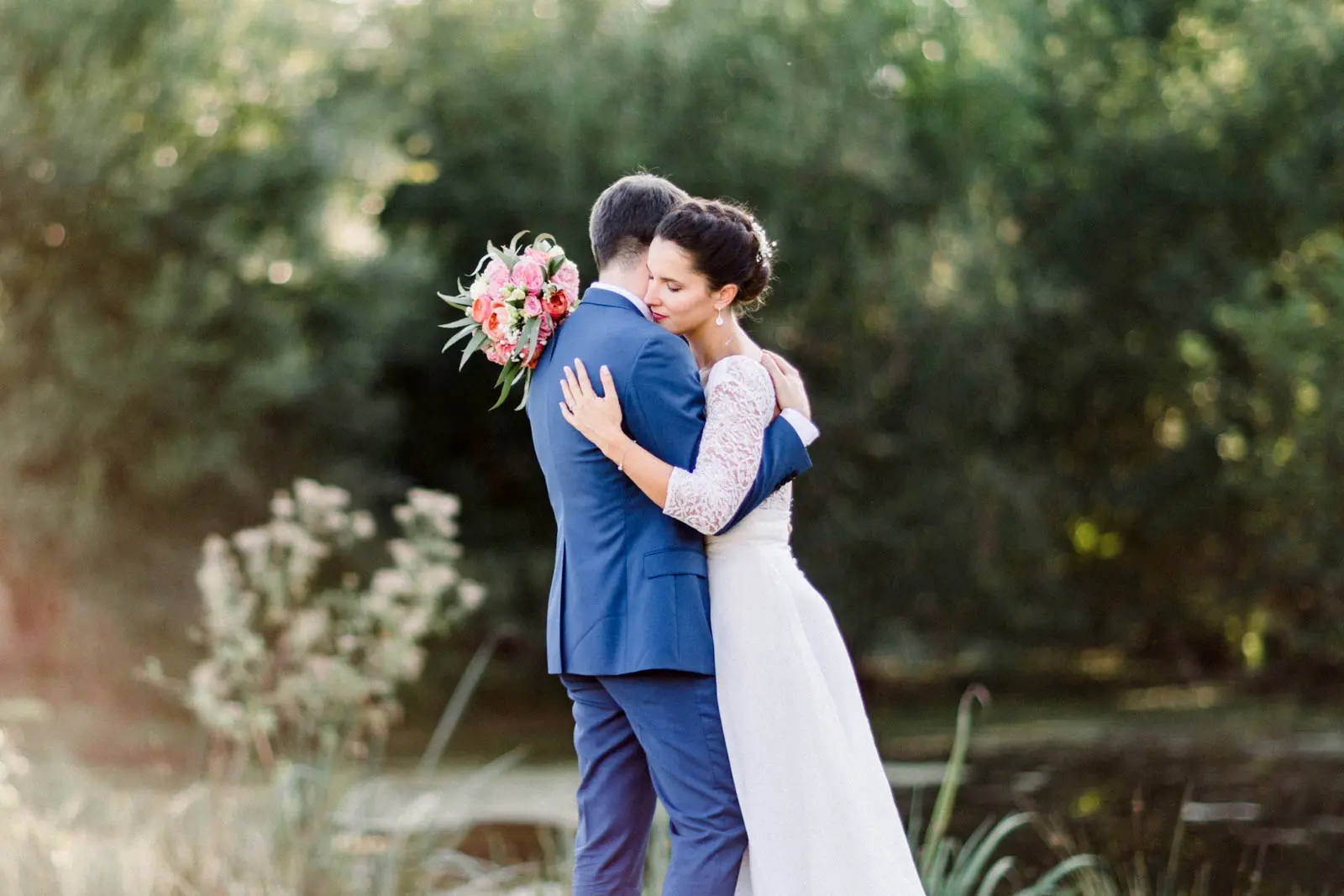Bride and groom embracing by wild grasses at the water's edge, landscape view