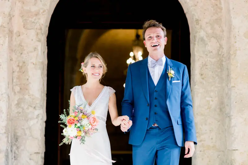 Bride and groom walking out hand in hand beneath a stone archway