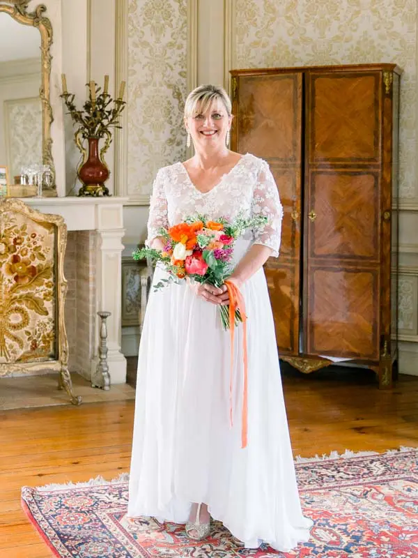 Hélène in a lace gown holding her coral bouquet in a château salon
