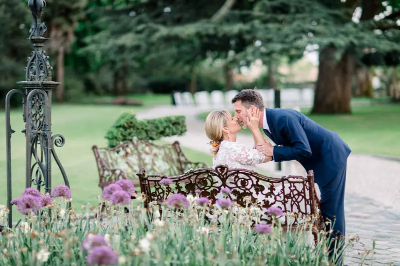 Groom kissing the bride seated on a wrought-iron bench among flowers