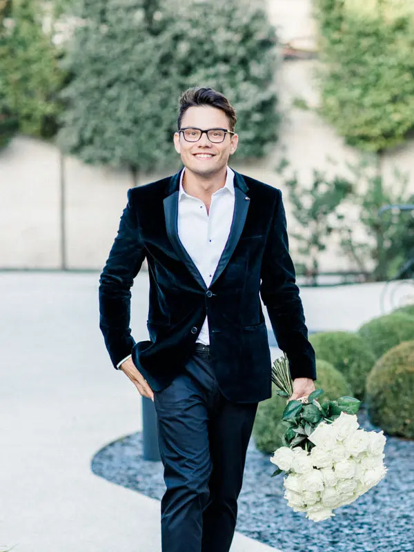 A man in a velvet suit walking with a bouquet of white roses