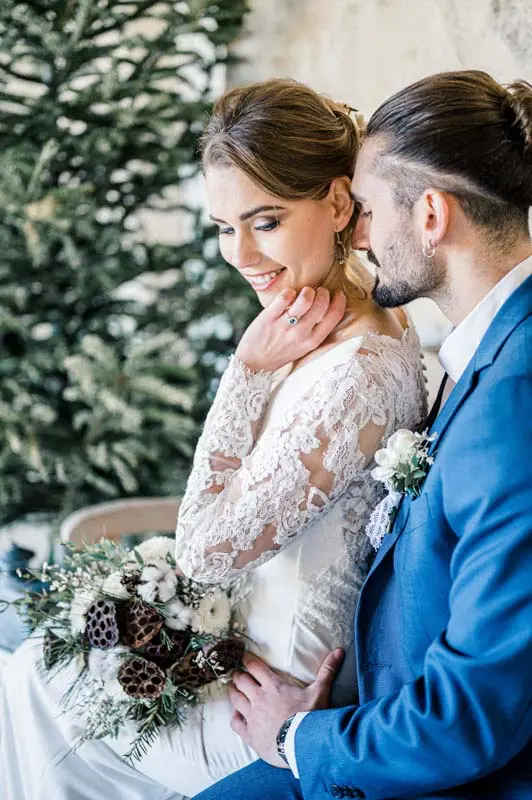 Bride in lace smiling, cheek against the groom, in front of fir trees