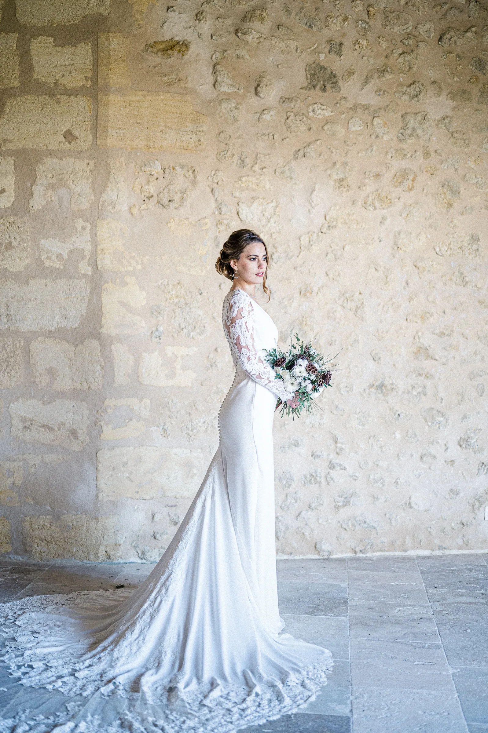 La mariée en robe à traîne devant un mur de pierre blonde