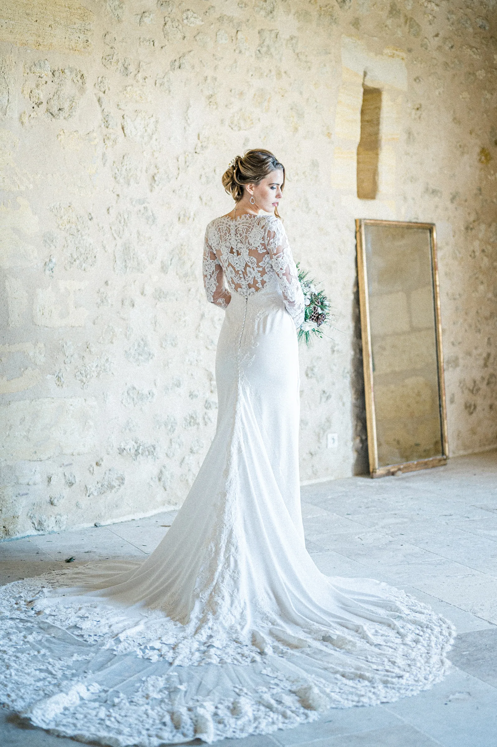 La mariée de dos dévoile sa longue traîne en dentelle dans la salle en pierre