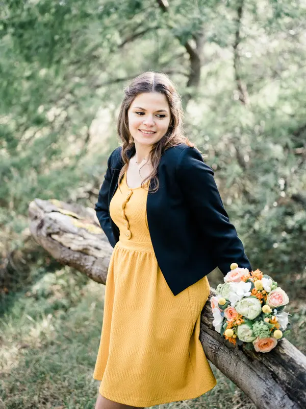 The bride-to-be in a yellow dress and black jacket leaning on a log