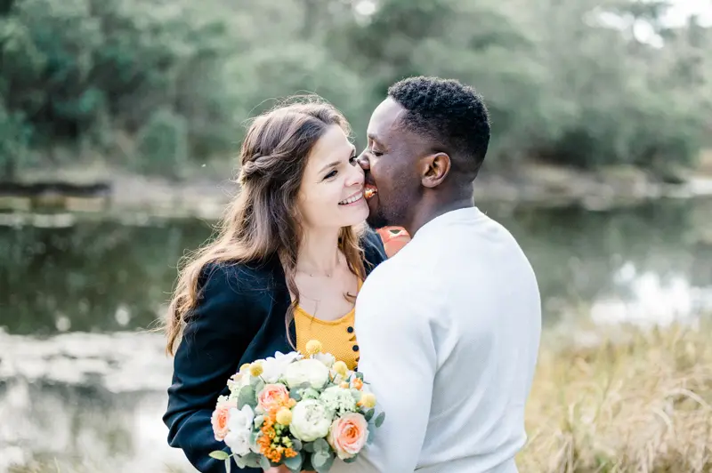 He kisses her cheek as they stand by the water