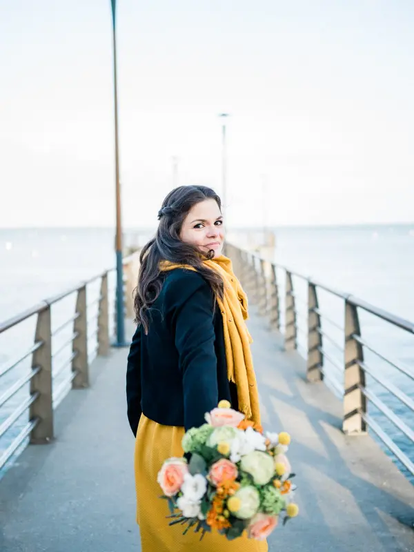 The bride-to-be in a yellow scarf walking along the jetty with her bouquet