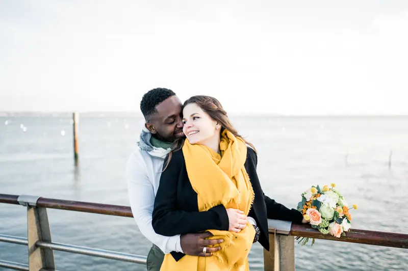 The couple embracing against the jetty railing, facing the bay
