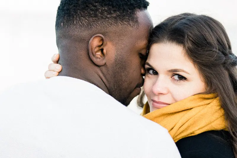 She looks into the lens over her partner's shoulder as he hugs her