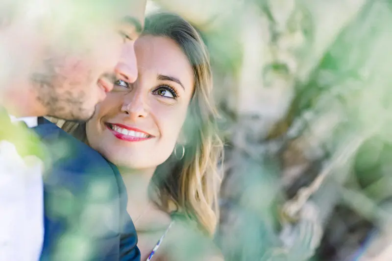 The bride smiling as the groom kisses her, seen through the leaves