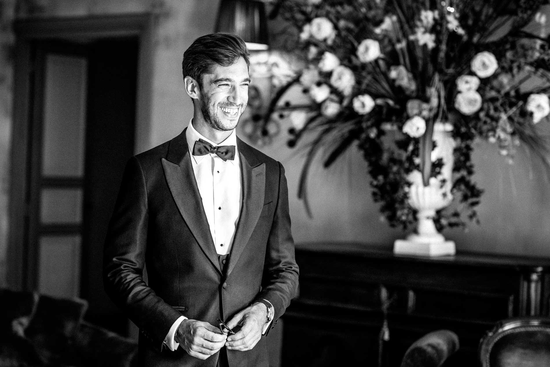 The groom in a tuxedo smiling beside a floral arrangement, in black and white