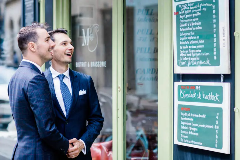 The two grooms smiling outside a café window in central Bordeaux
