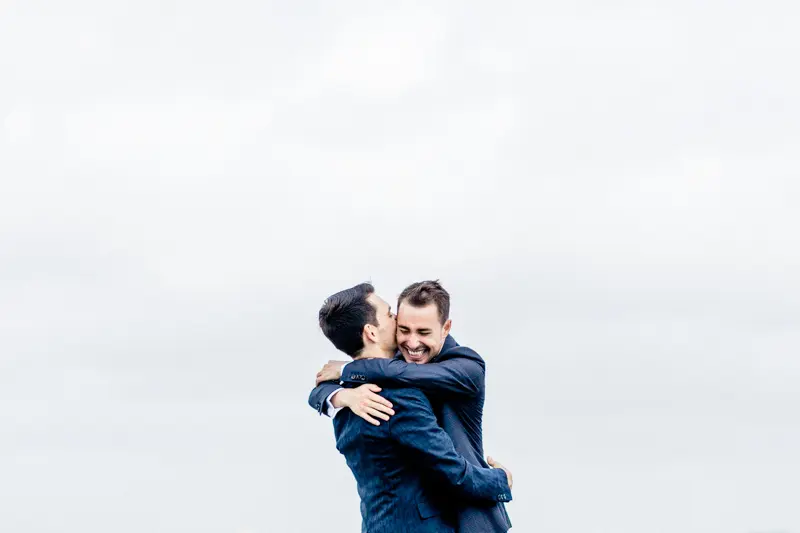 One groom leaping into the other's arms against a white sky in Bordeaux
