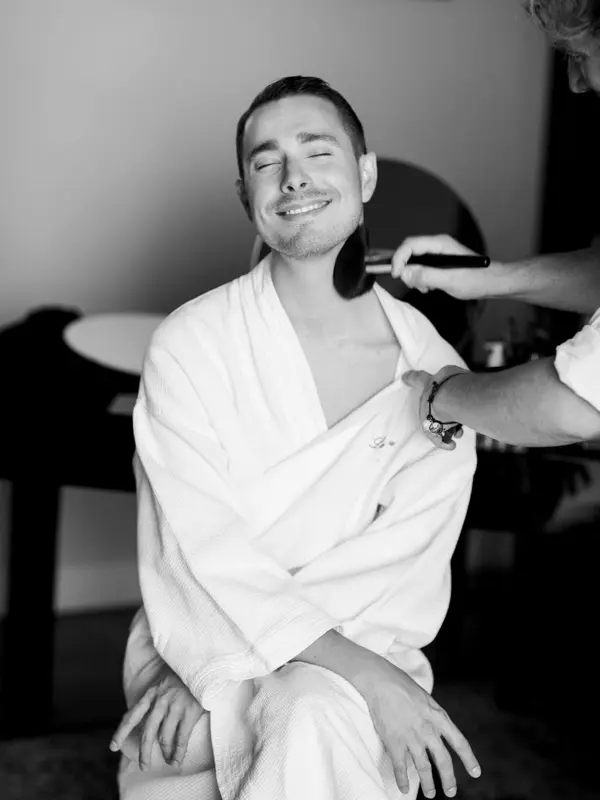 A groom in a bathrobe having his make-up done while getting ready, in black and white