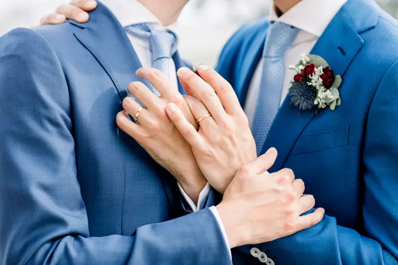 The grooms' hands and wedding rings on blue suits with buttonholes