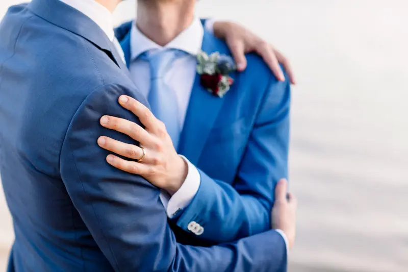 A hand resting on a groom's blue-suited shoulder with a burgundy buttonhole