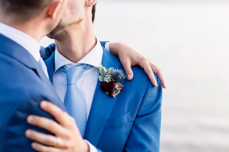 The grooms cheek to cheek by the water, detail of the blue suits
