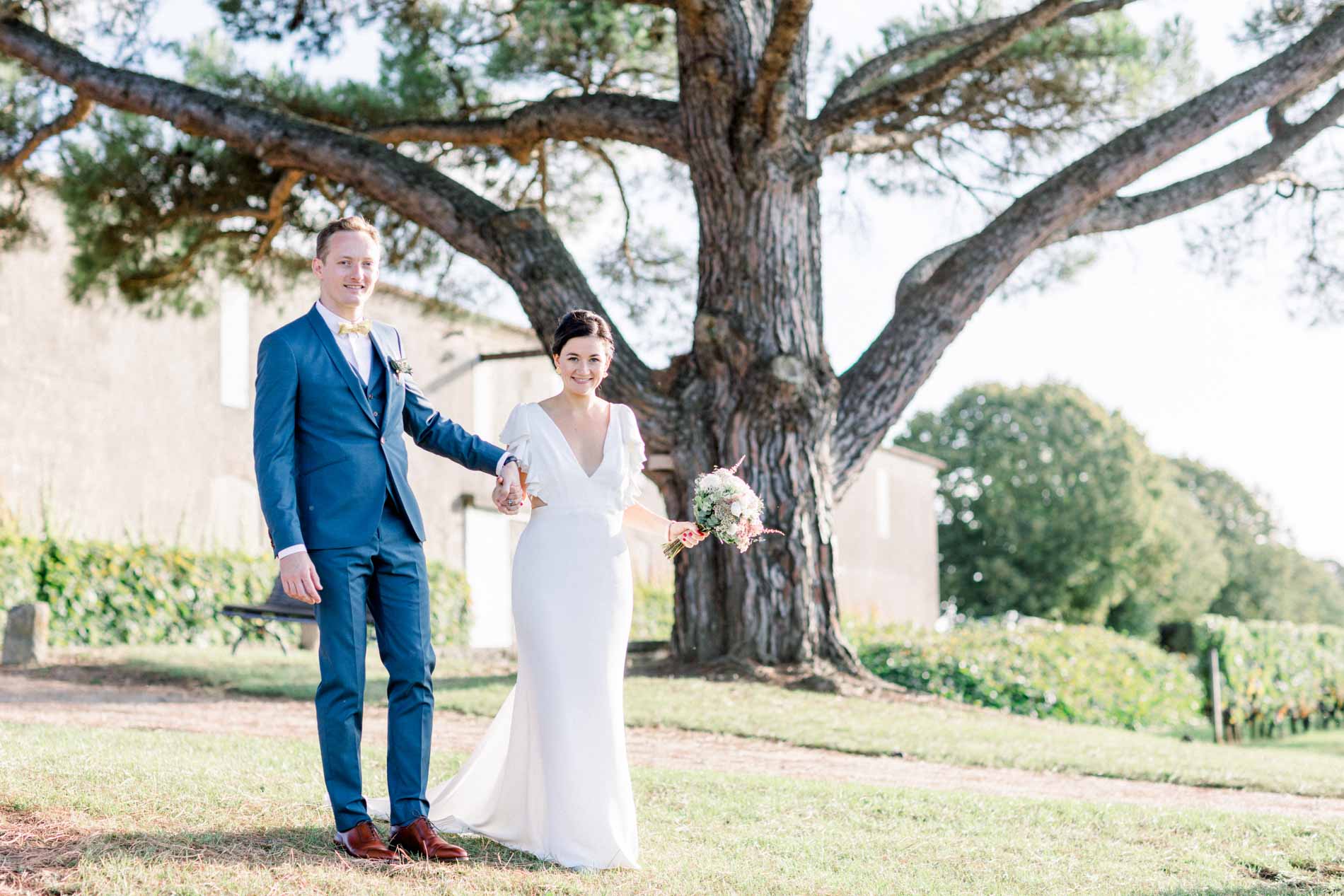 The bride and groom hand in hand under a large tree in the château grounds