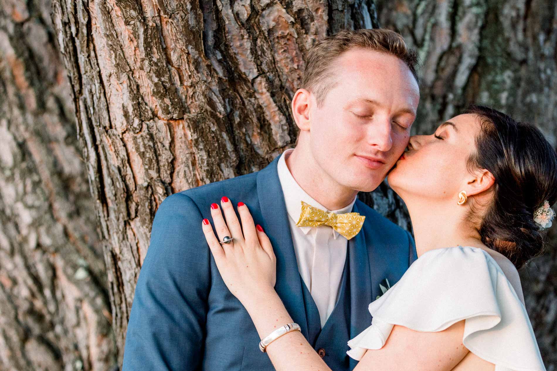 The bride kissing the groom's cheek as he closes his eyes against the tree