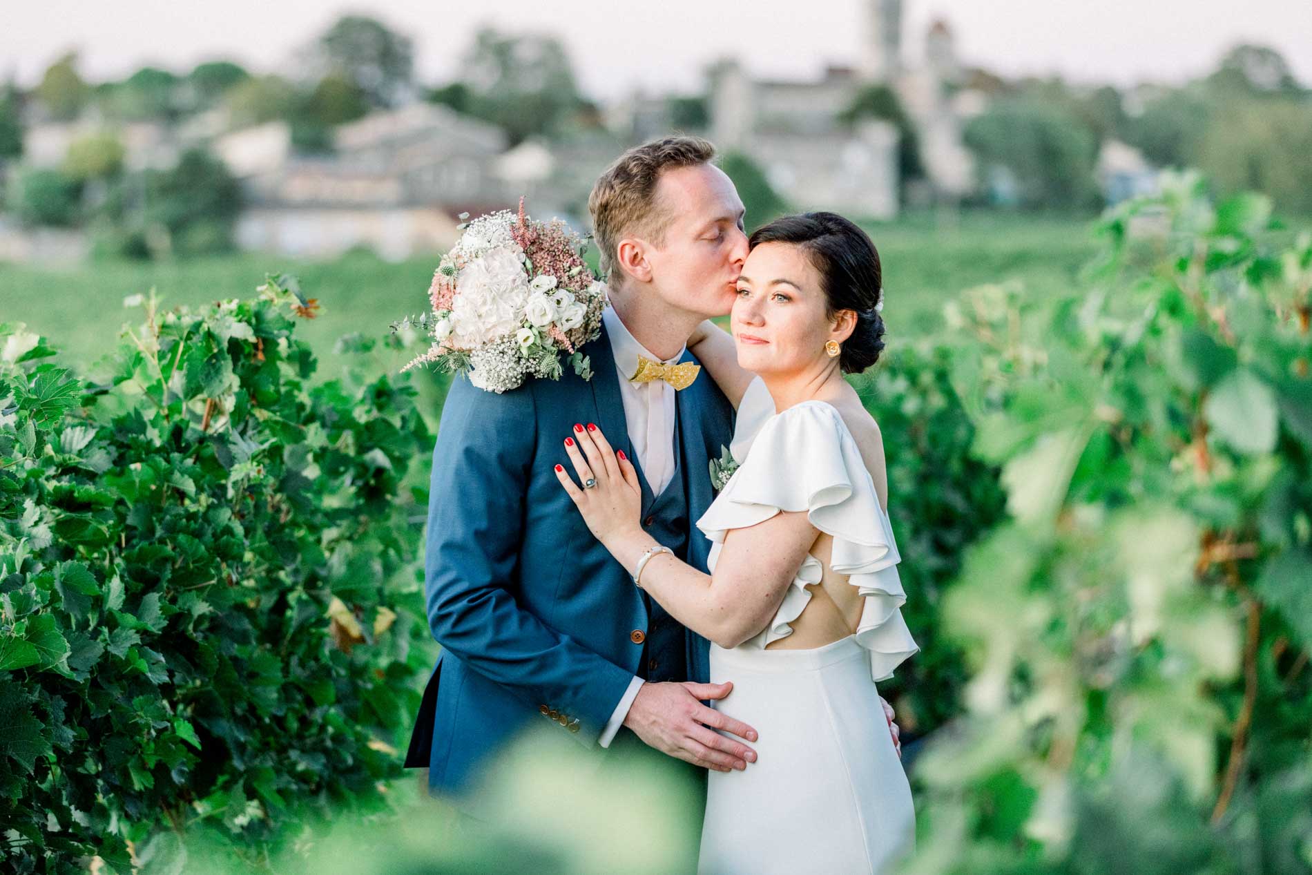 The groom kissing the bride's forehead between the rows of vines