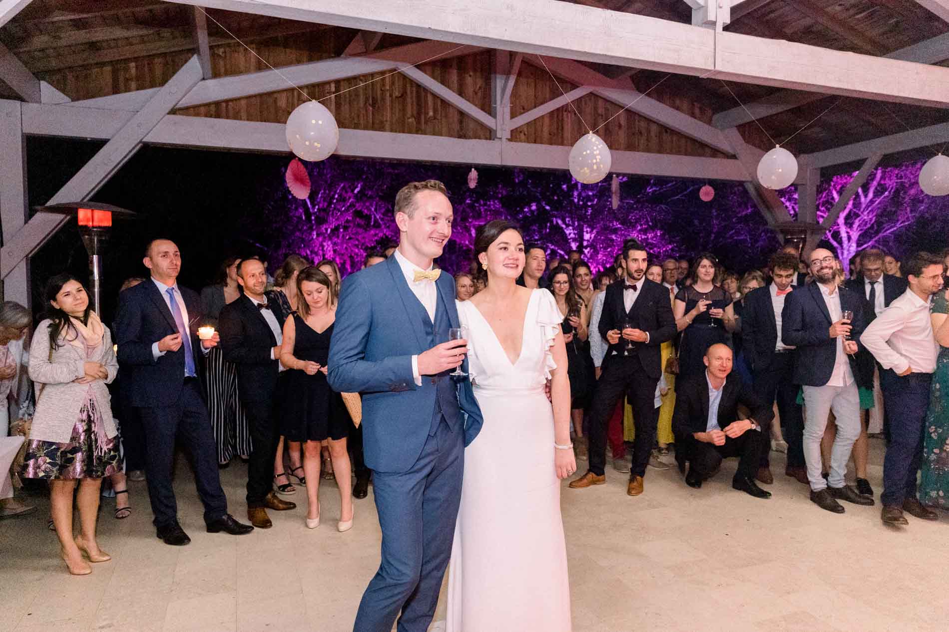 The bride and groom surrounded by guests beneath purple-lit beams