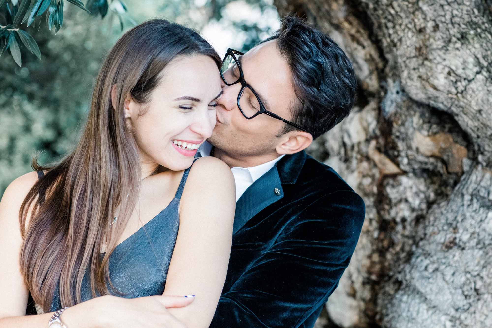 The man kissing his laughing partner's cheek against an old tree trunk