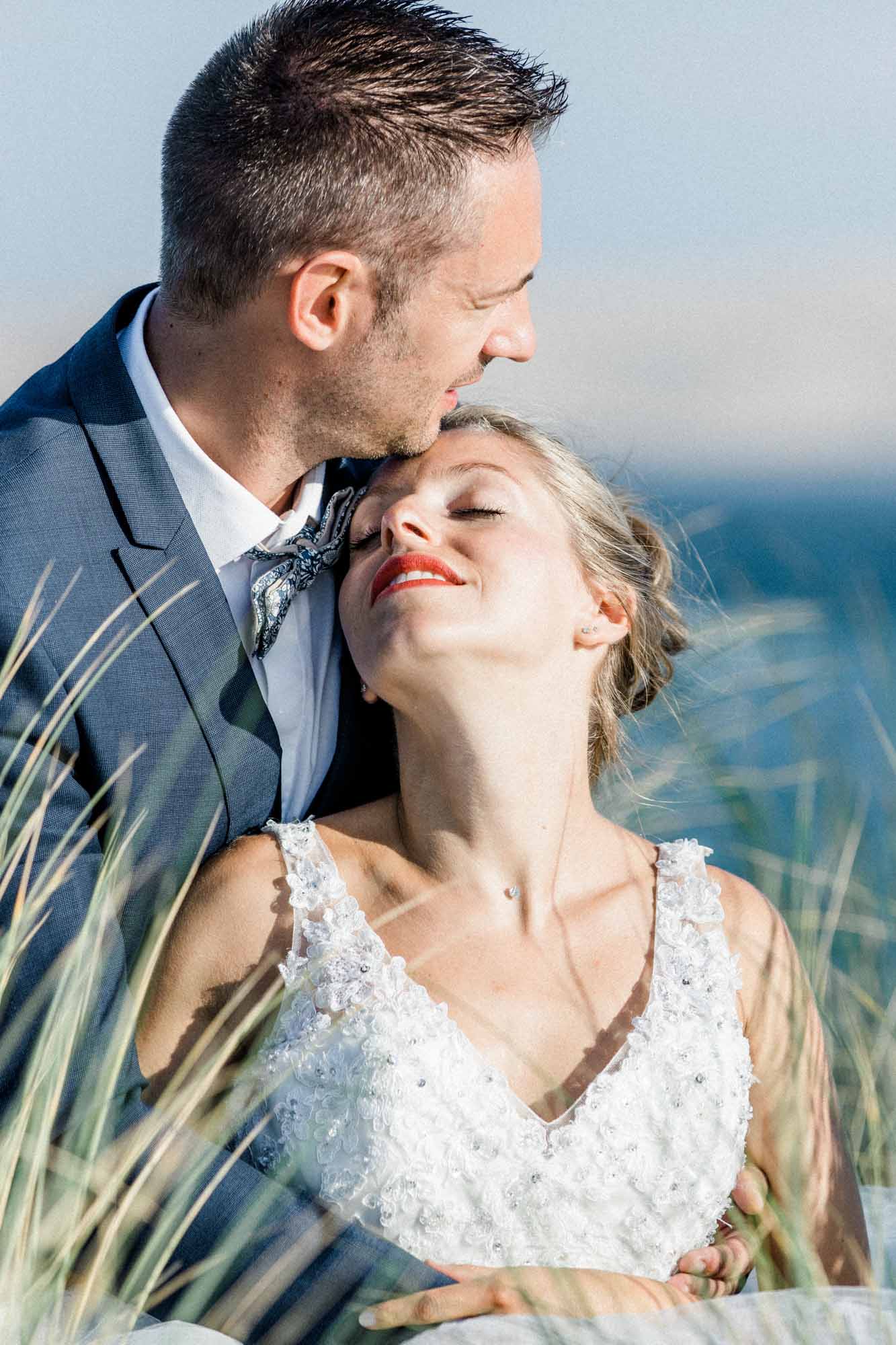 Le marié embrasse le front de la mariée, yeux fermés, dans les dunes