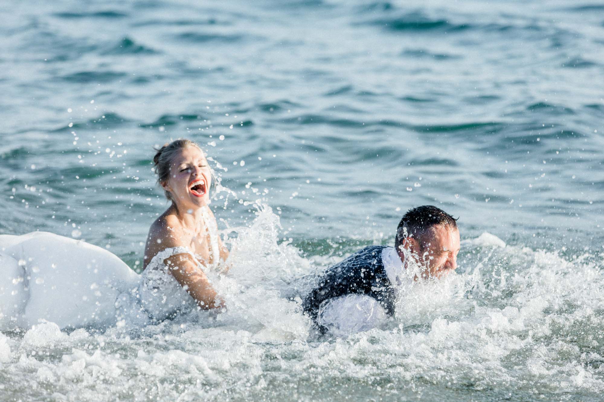 Les mariés éclaboussés en riant dans les vagues, robe trempée