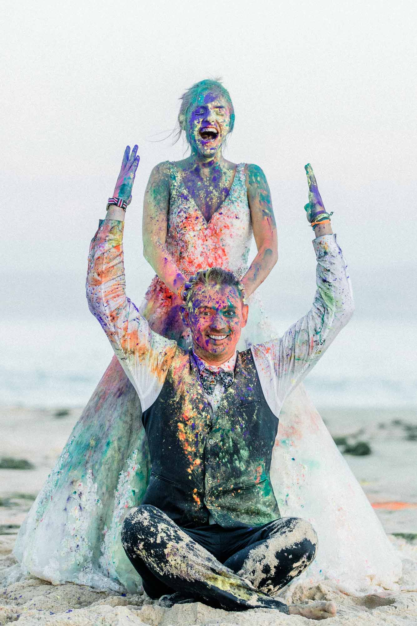 La mariée lève les bras derrière le marié, tous deux couverts de couleurs