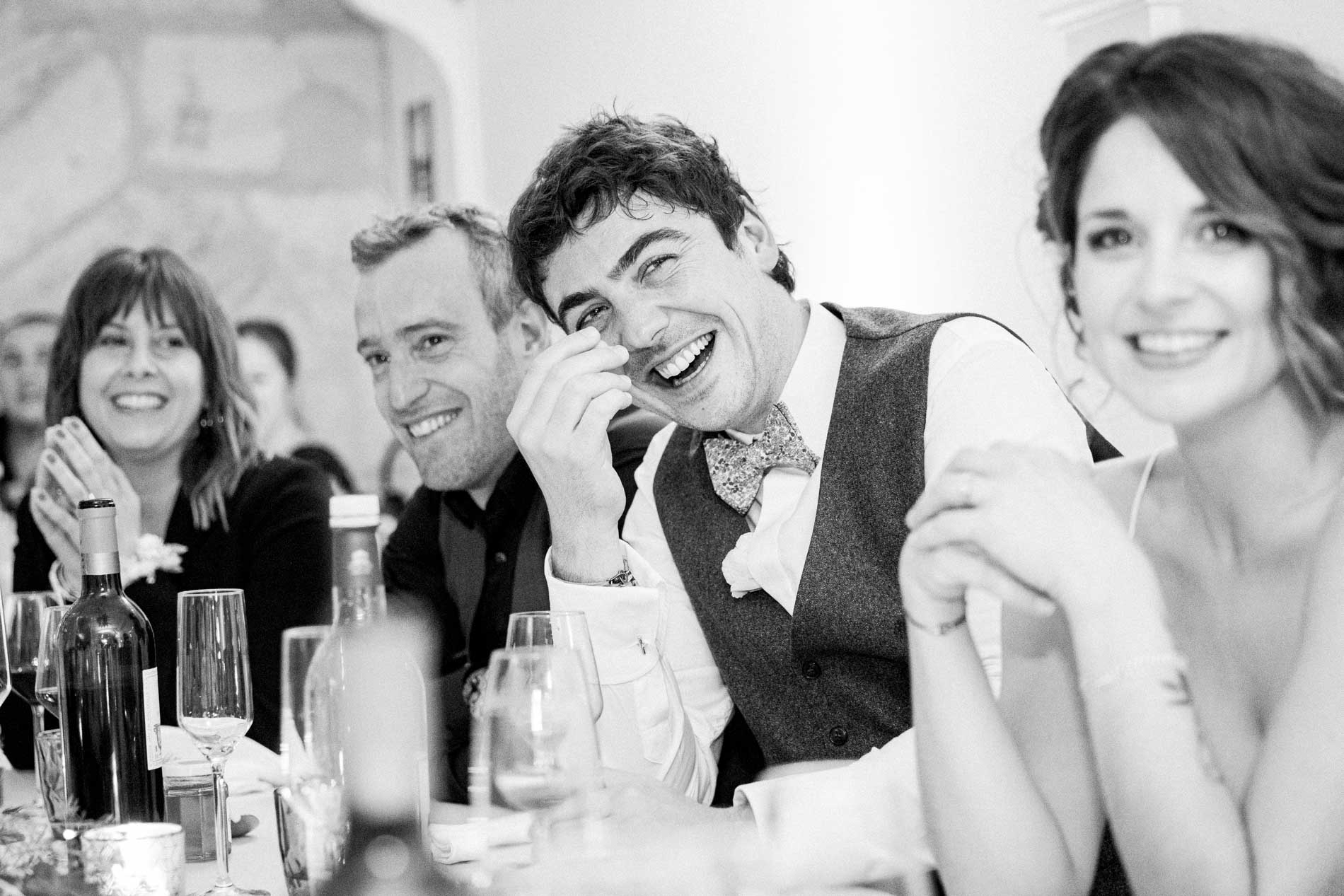 The groom laughing heartily at the table beside the bride, in black and white
