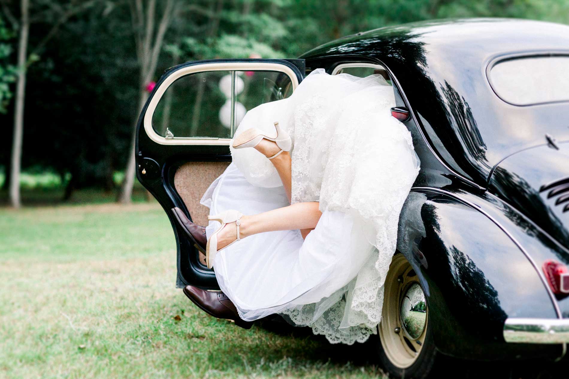 Bride laughing as she climbs into the black vintage car, her gown billowing