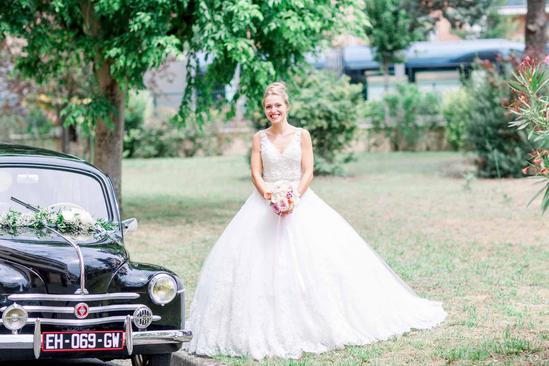 Bride posing in front of a black vintage car decorated with flowers in Mérignac