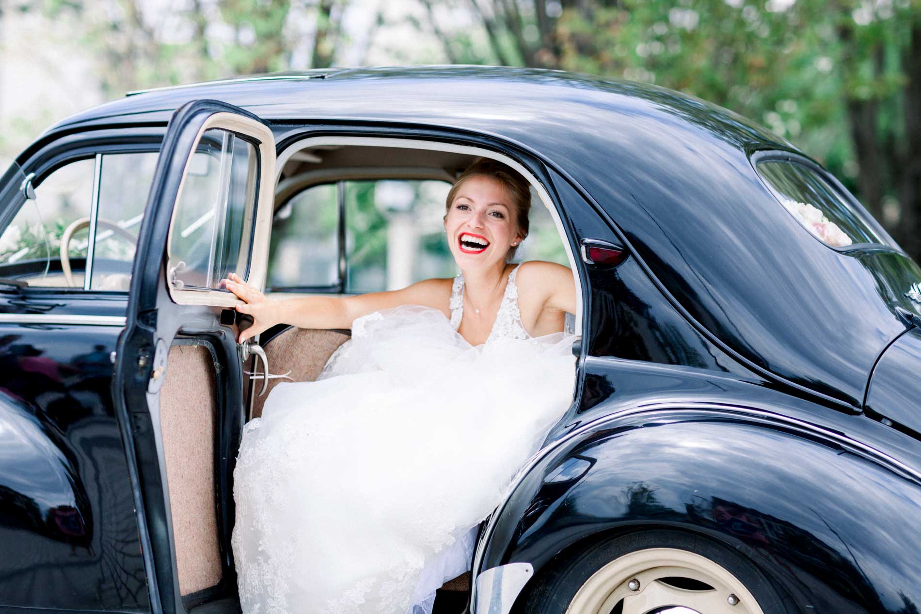 Laughing bride sitting in a black vintage car with the door open