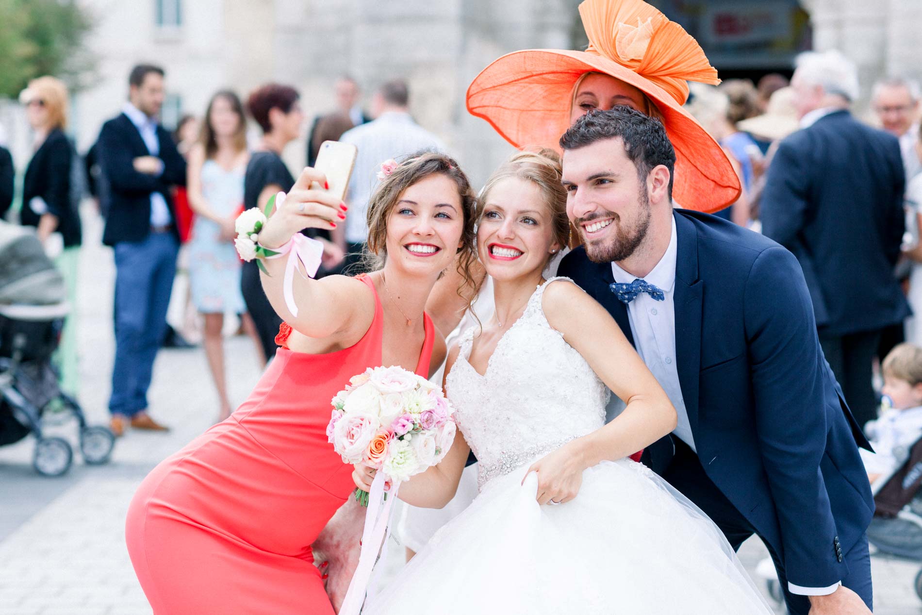 Newlyweds taking a selfie with a guest in an orange hat outside the Mérignac church