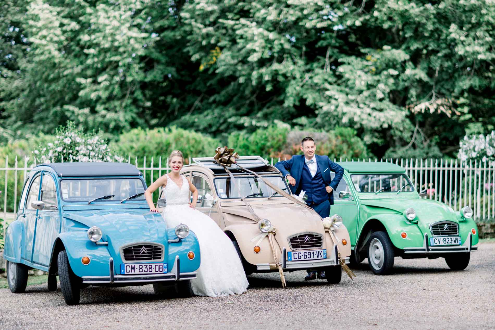 Bride and groom posing among three colourful Citroën 2CVs by the château gates