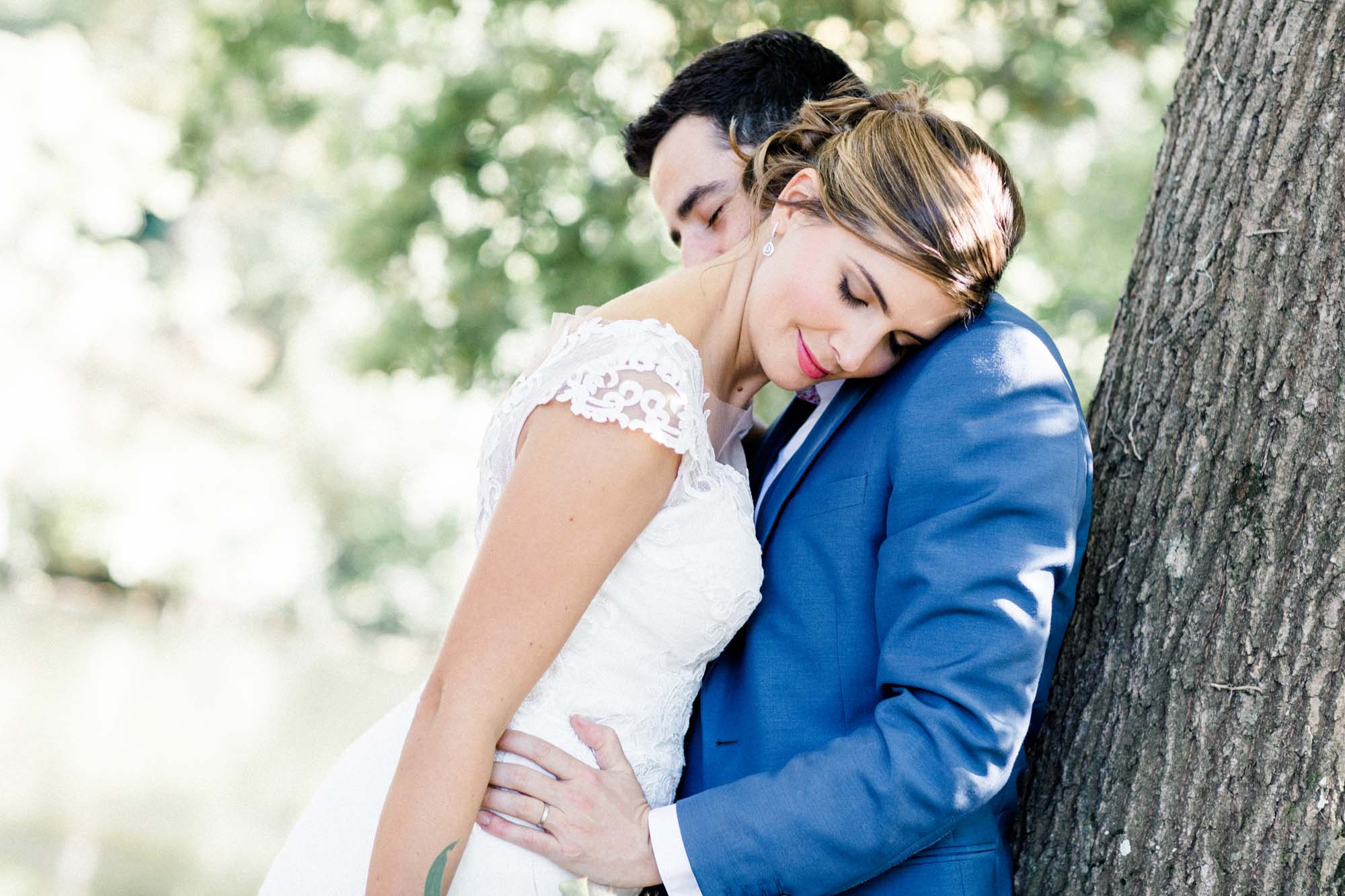 The bride nestled against her husband as he leans on a tree trunk