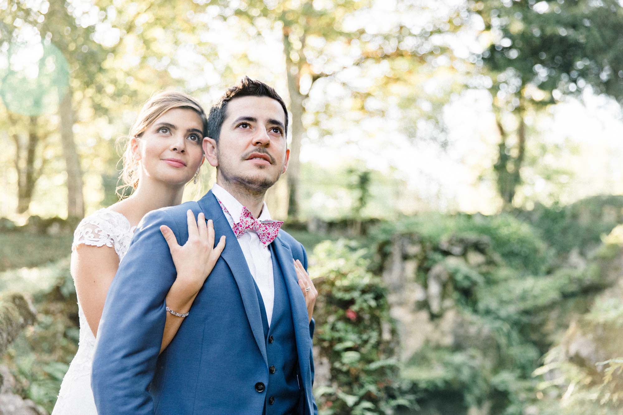 Groom in a pink bow tie embraced by the bride under the trees