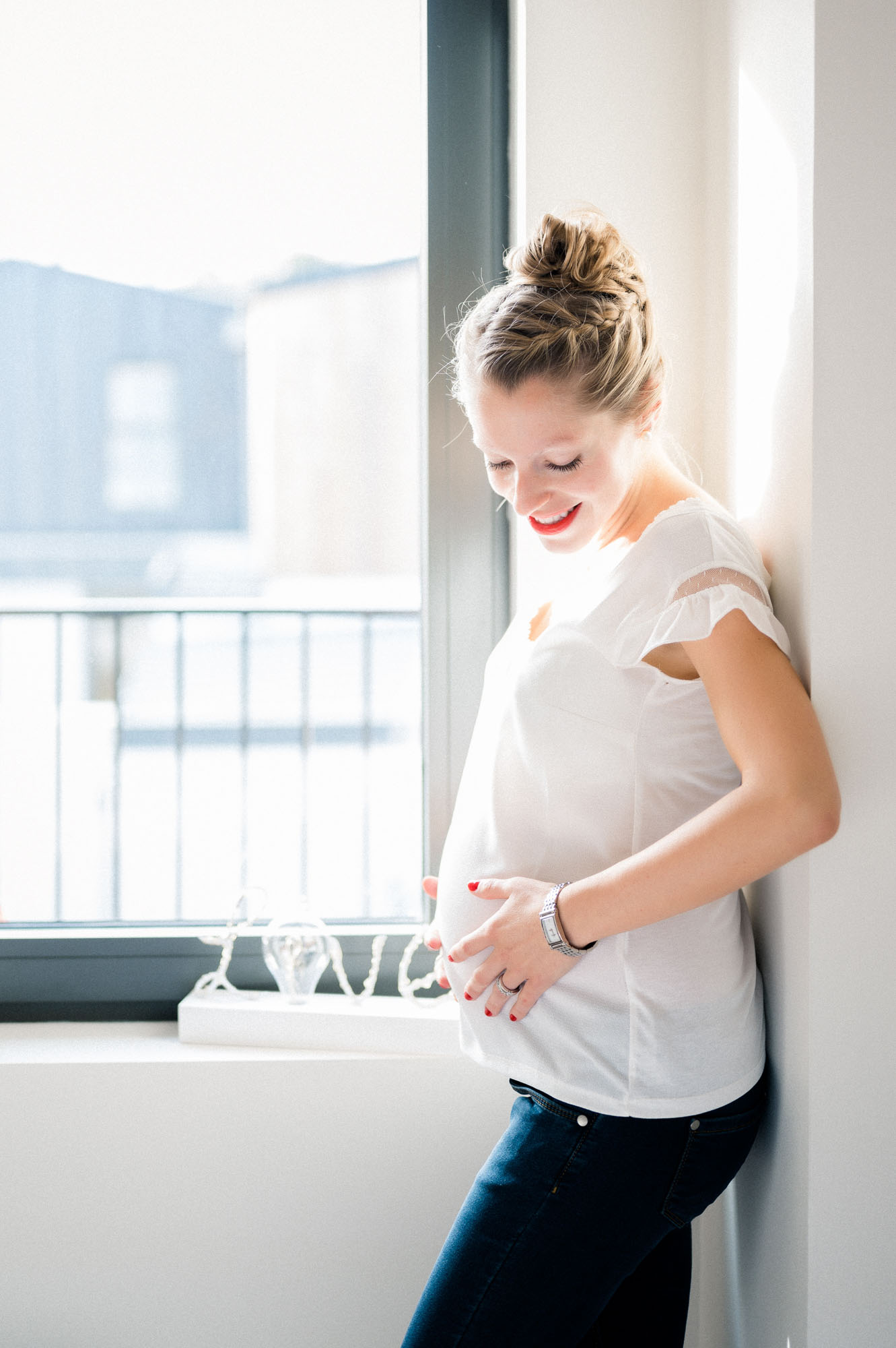 Smiling mum-to-be leaning by a window with her hands on her bump, in Mérignac