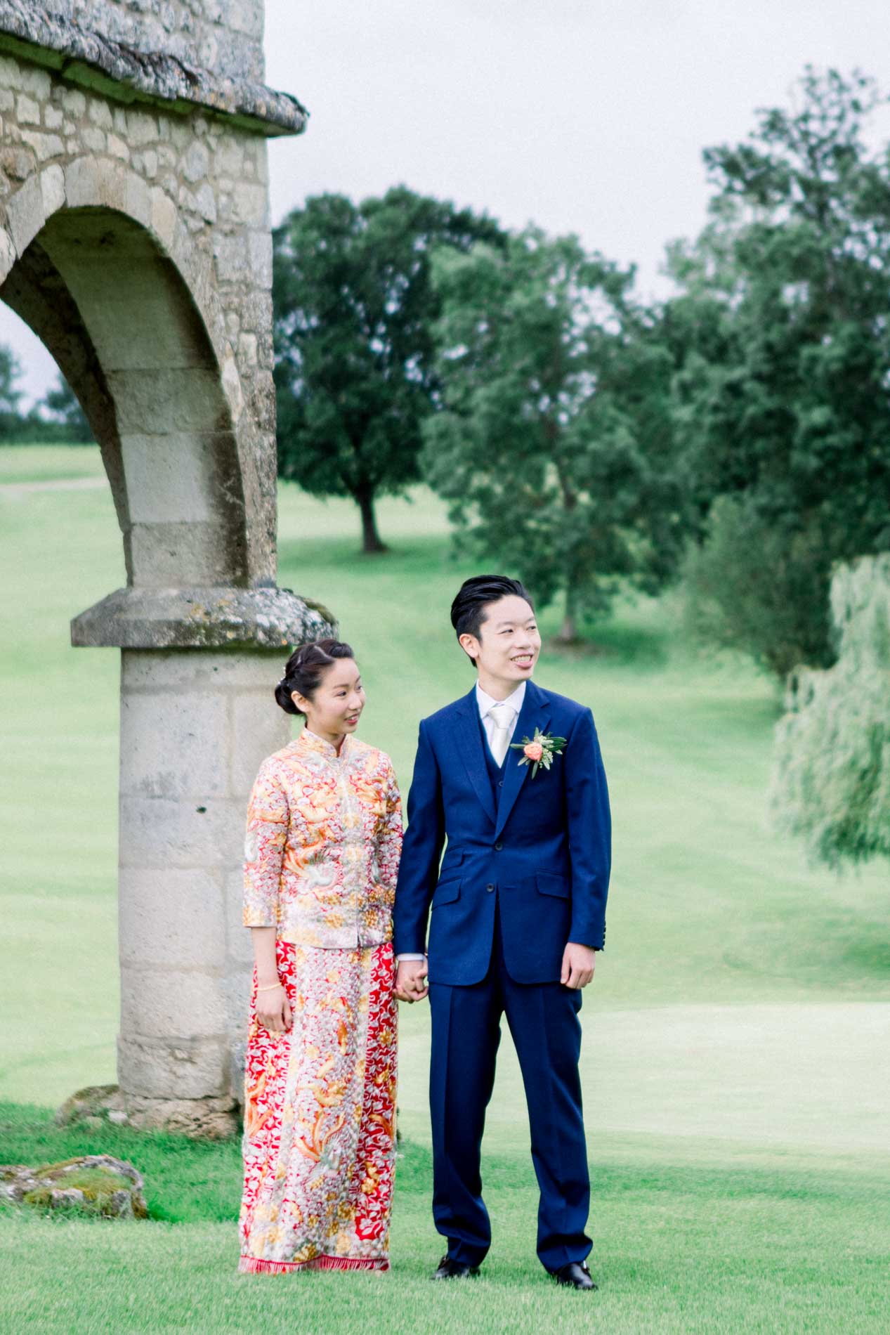 The couple posing by a stone arch in the park, bride in red traditional dress