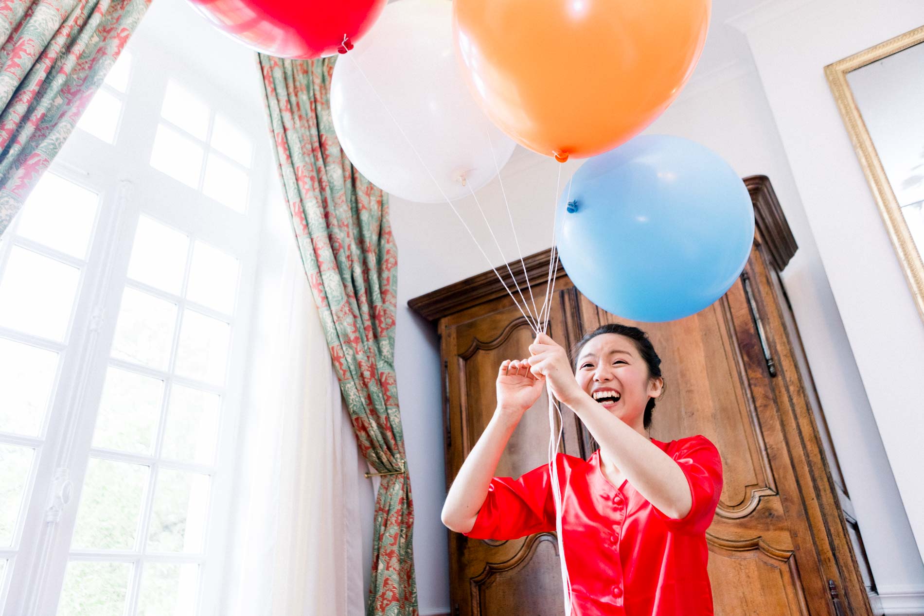 Young woman in red holding a bunch of colourful balloons in a bedroom