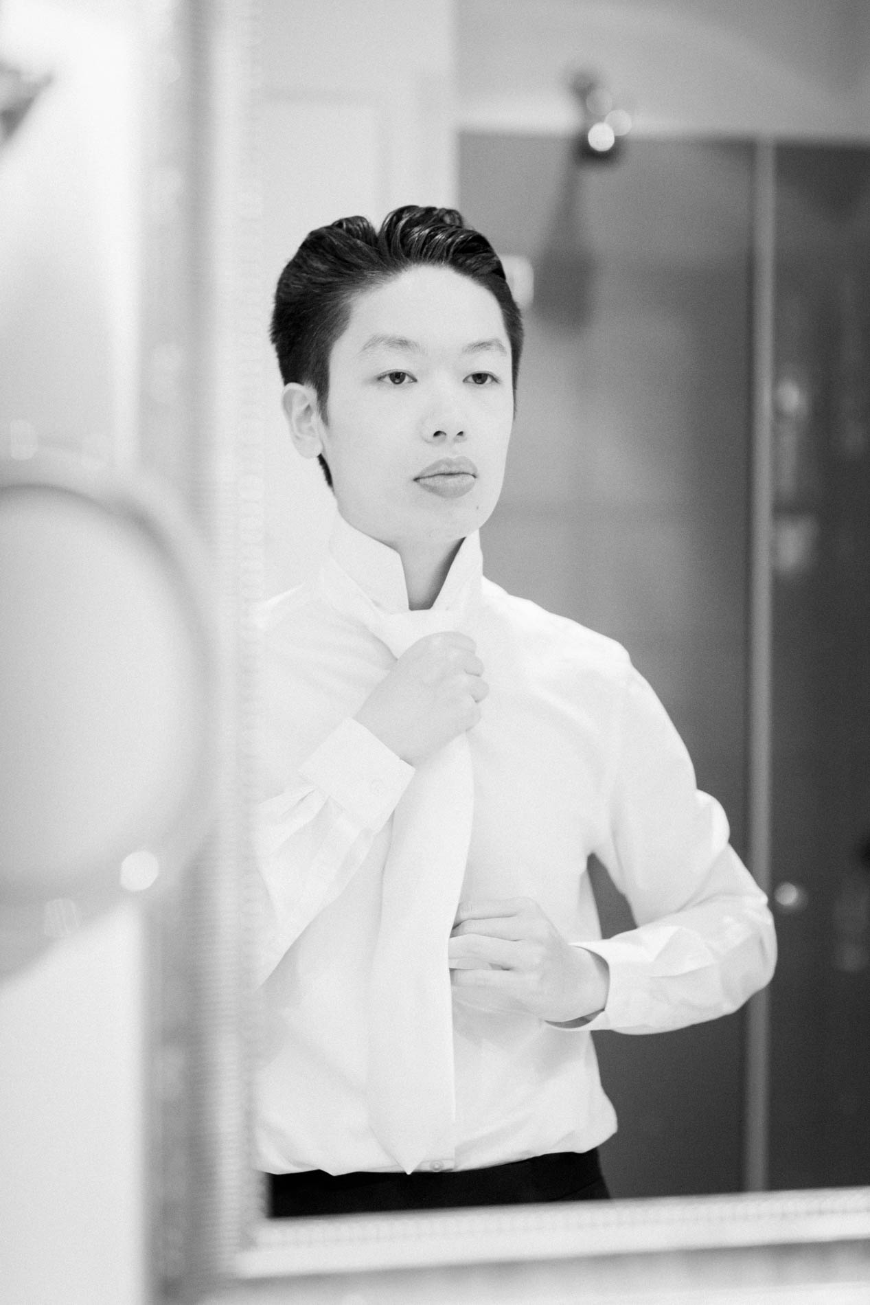 The groom tying his tie in front of the mirror, in black and white