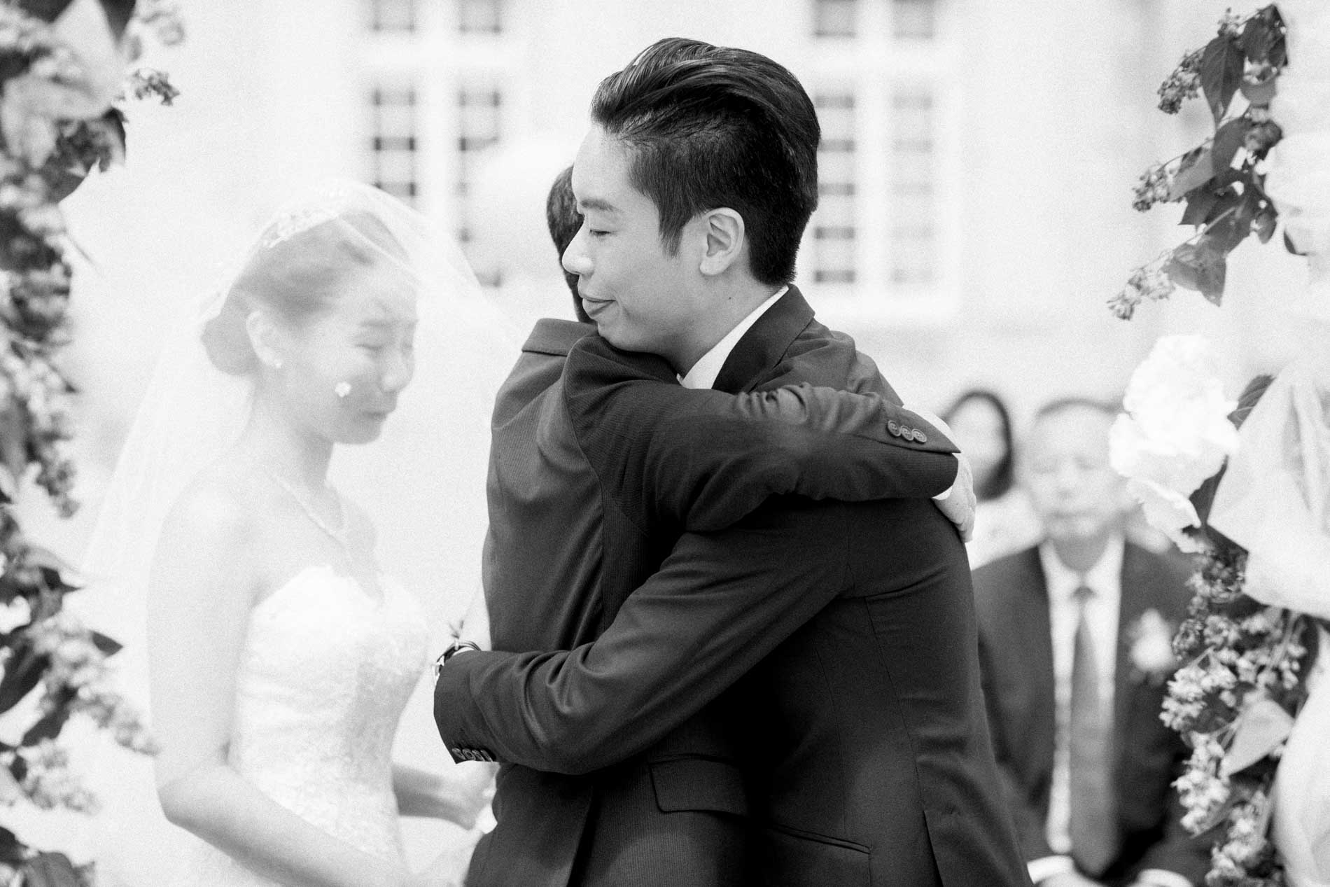 Emotional hug between the groom and a relative during the outdoor ceremony, in black and white