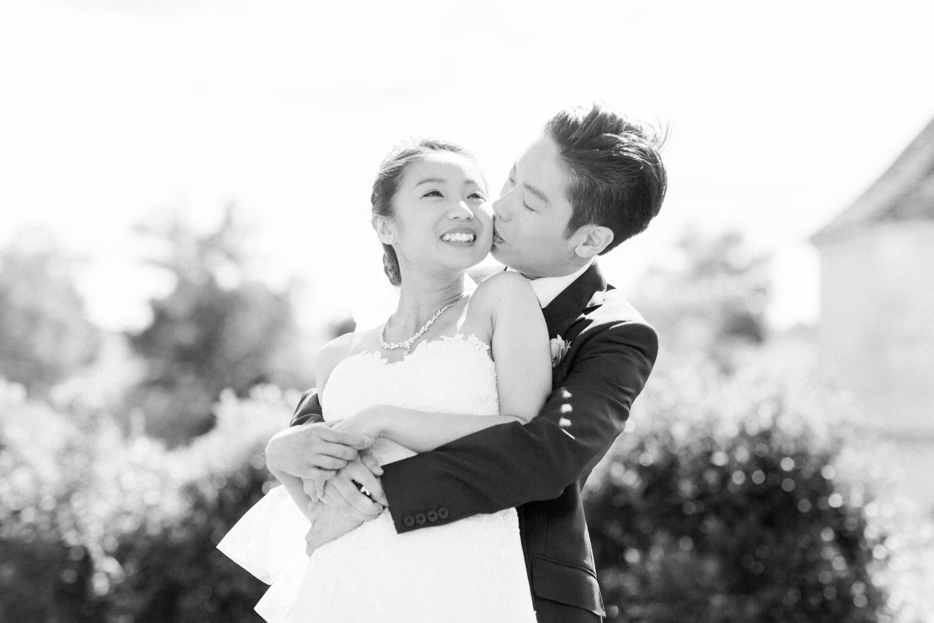 The groom kissing the bride's cheek as he hugs her, in black and white